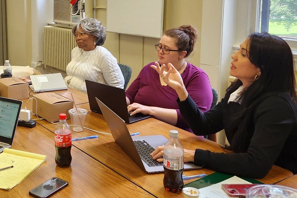 A group of colleagues sits around a conference table during a meeting as a woman in a black jacket gestures while speaking. Several participants work on laptops, and notebooks, drinks, and other materials are spread across the table.
