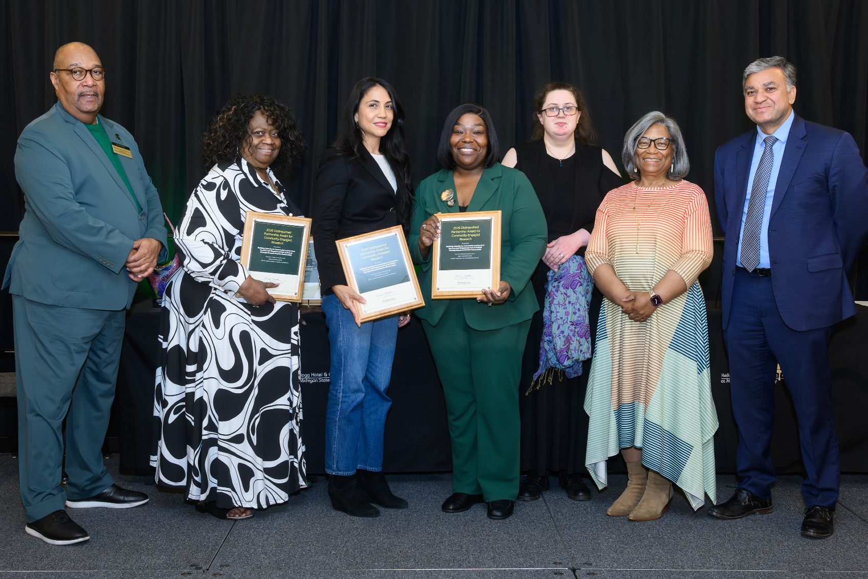 A group of seven people stands together at an awards ceremony, with three recipients holding framed plaques while the others pose alongside them.