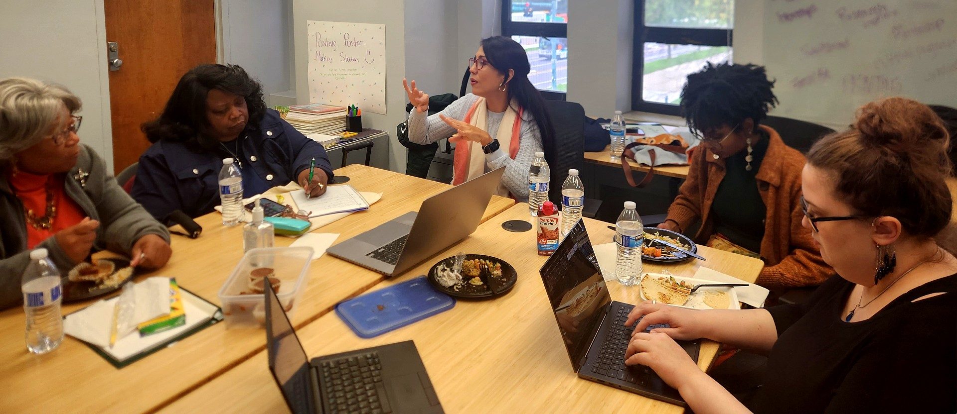 A group of people sits around a conference table during a meeting. One woman gestures while speaking as others take notes, work on laptops, and listen.