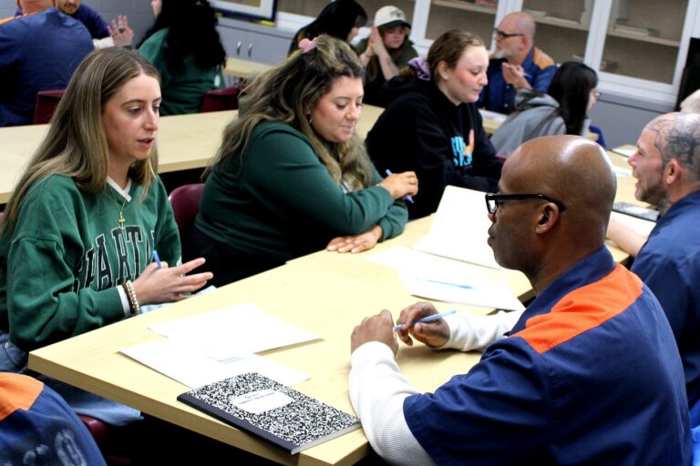Michigan State University students sit across from participants in blue-and-orange uniforms, talking and working together on a writing activity at classroom tables.