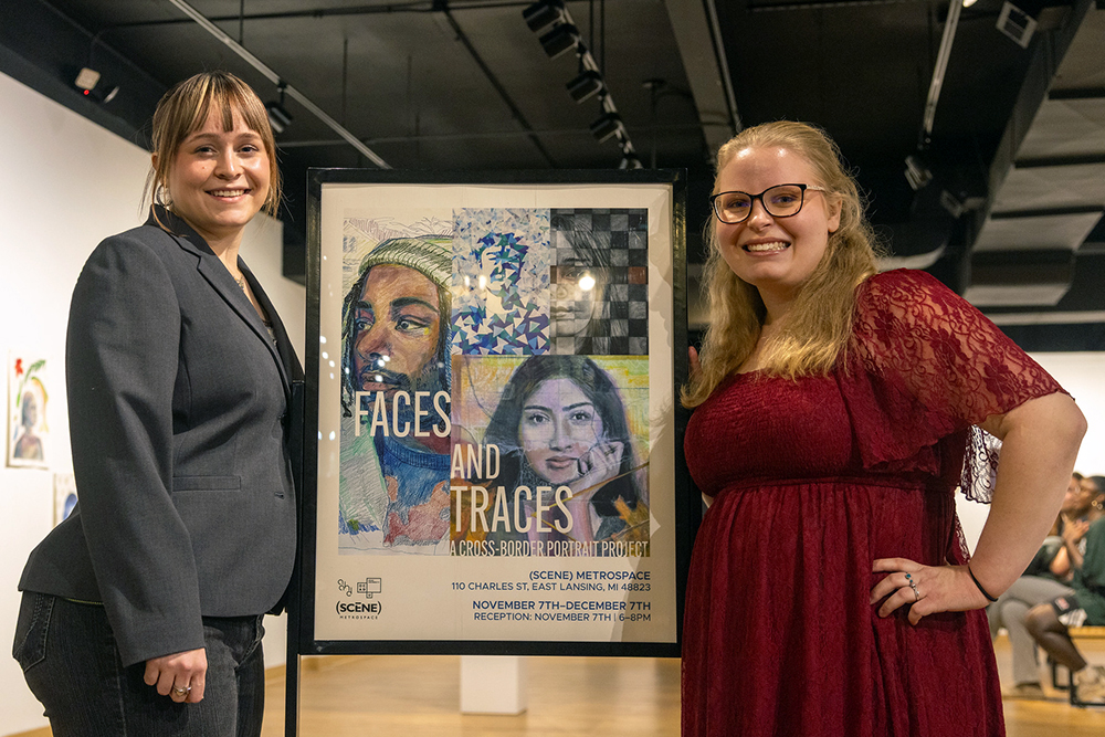 Two students stand on either side of a sign that has portrait drawings and the words: “Faces and Traces: A Cross-Border Portrait Project” as the exhibition title held at (SCENE) Metrospace in East Lansing. Behind the students is a gallery setting with people viewing art on the walls.