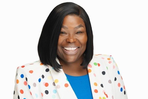 Studio portrait of a smiling woman with shoulder-length black hair wearing a white blazer with colorful polka dots over a bright blue top against a white background.