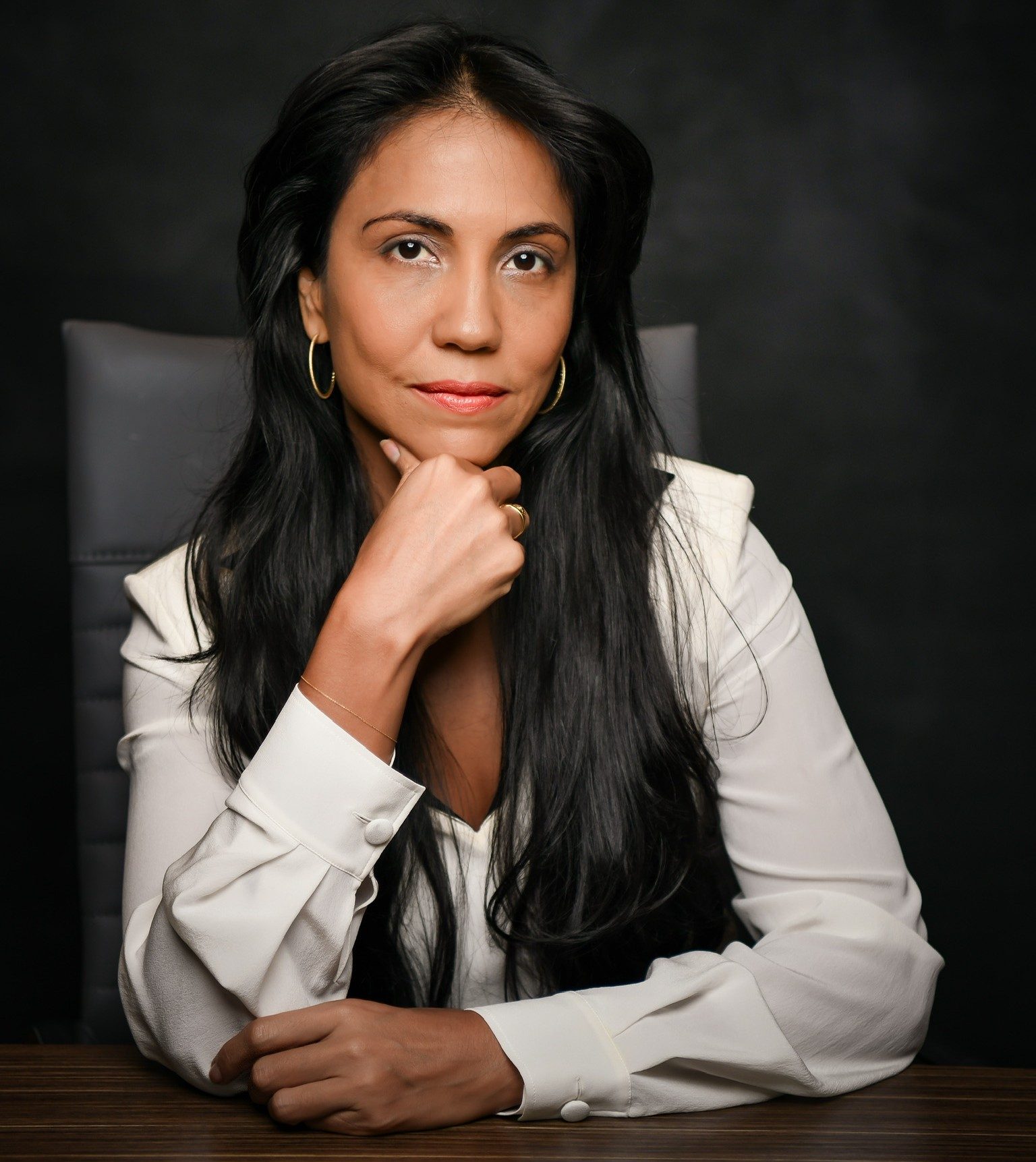Studio portrait of a woman with long dark hair seated at a desk with her chin resting on one hand. She is wearing a white blouse and gold hoop earrings against a dark background.