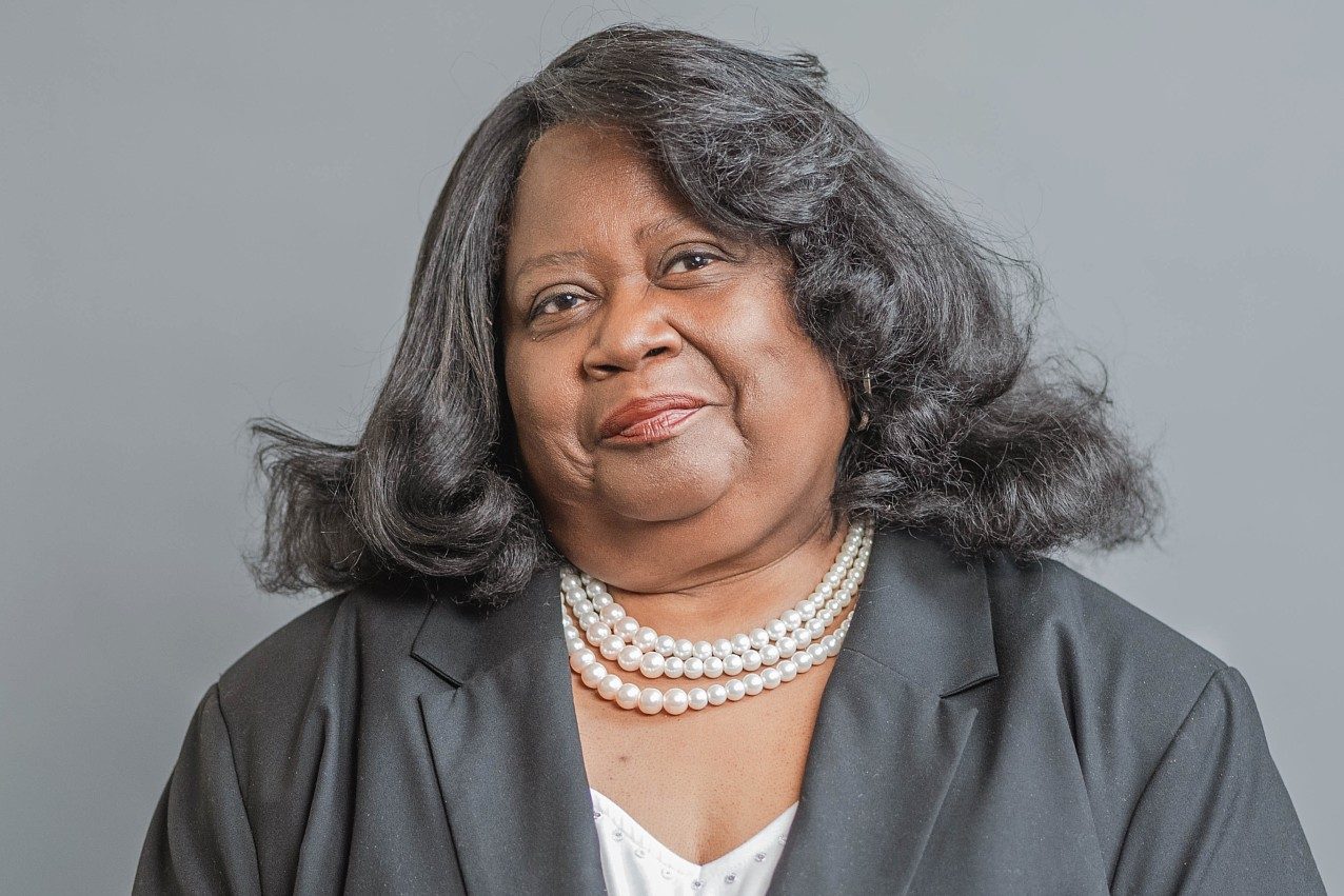 Studio portrait of a woman with shoulder-length dark hair wearing a black blazer, a white blouse, and a layered pearl necklace against a gray background.