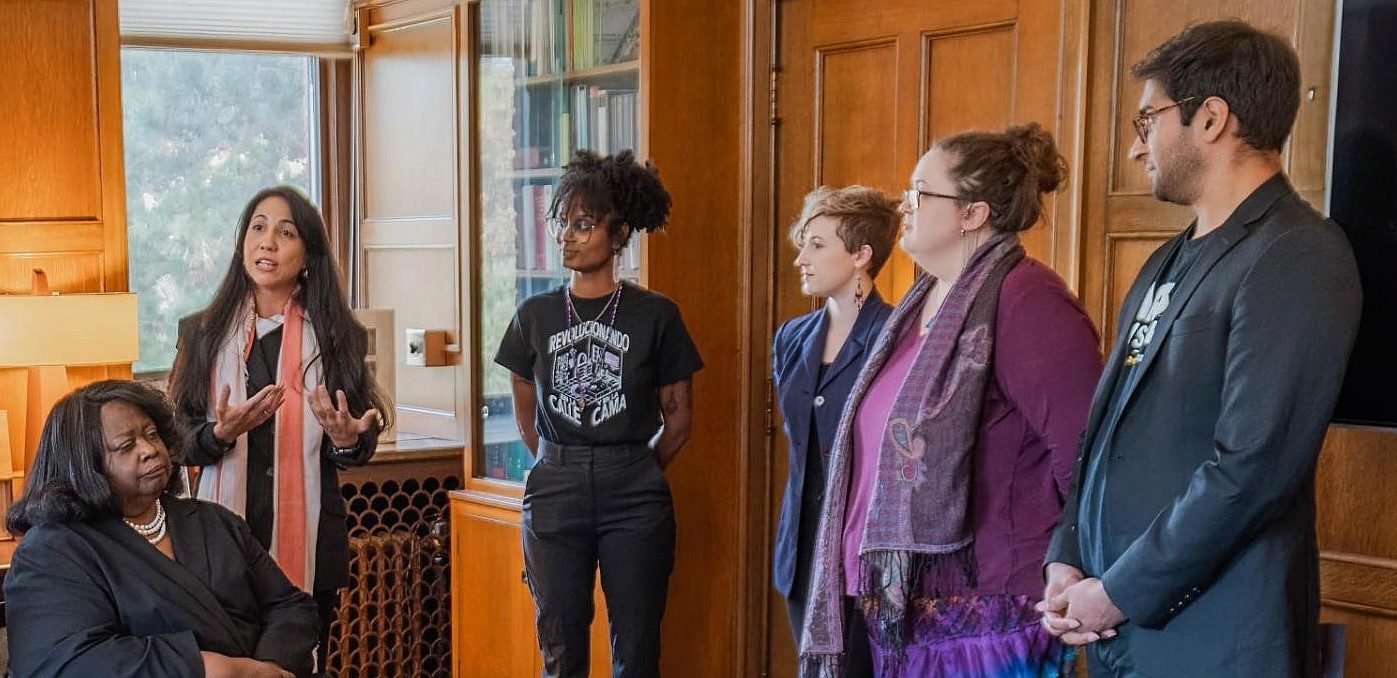 A woman speaks to a small group gathered in a wood-paneled room lined with bookshelves while another woman sits nearby listening. Four attendees stand facing the speaker. 