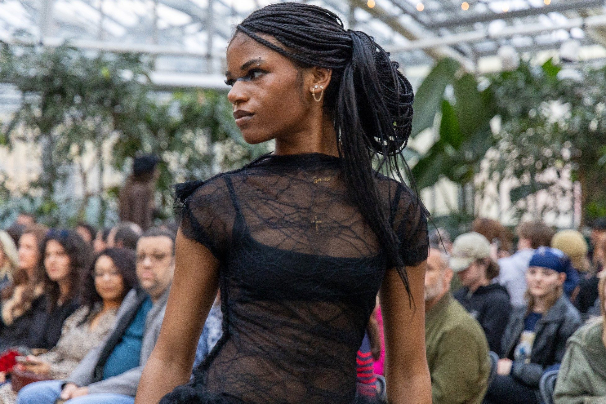 A close-up of a student model wearing a sheer black textured dress as she turns her head while walking the runway during Michigan State University's Studio 450 fashion show inside the Horticulture Gardens greenhouse. The audience and greenhouse plants are softly blurred in the background.