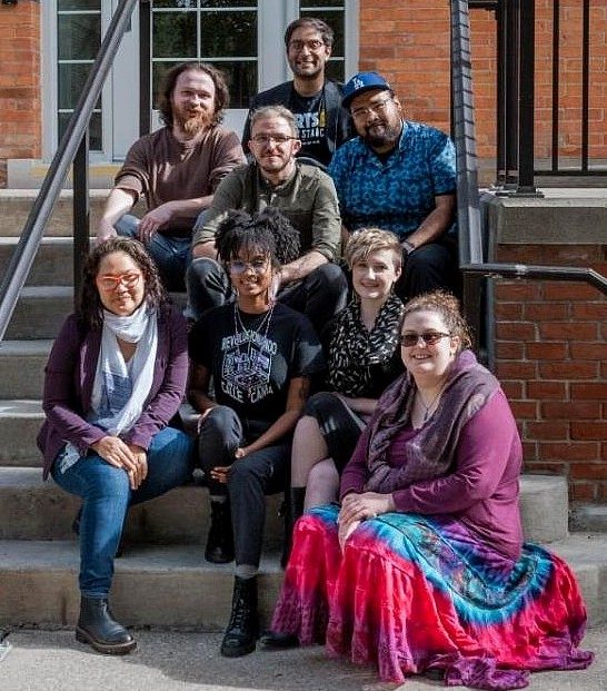 Eight people pose together on the front steps of a brick building on a sunny day. 