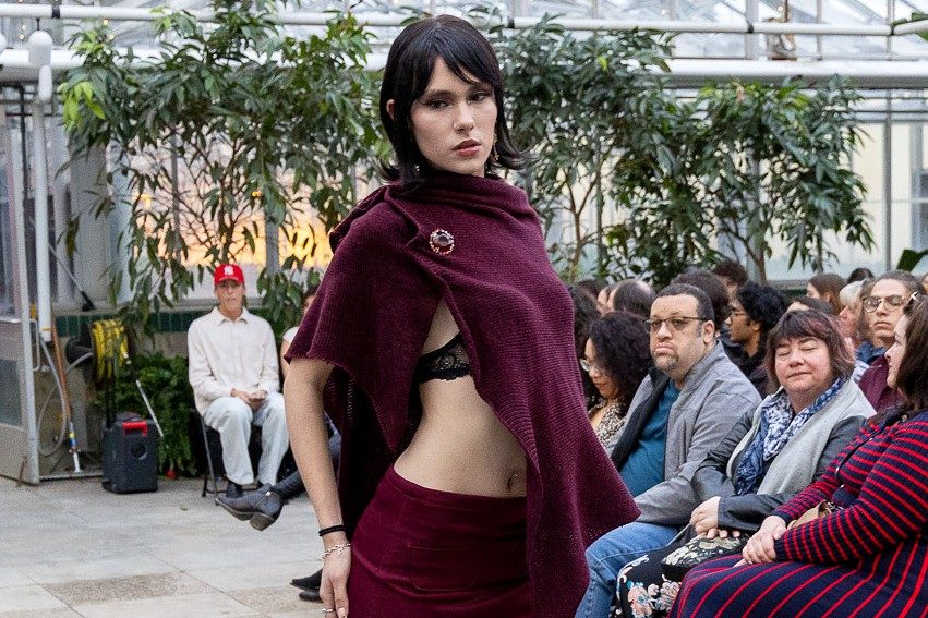 A student model poses on the runway inside Michigan State University's Horticulture Gardens greenhouse wearing a burgundy ensemble featuring a draped shawl-style top, and matching mini skirt during the Studio 450 fashion show. Audience members watch from both sides of the runway.