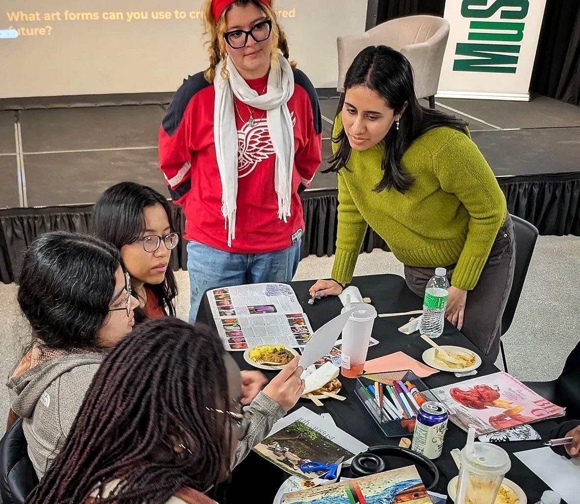 Aditi Gonuguntala leans over a table to talk with a group of students during a collaborative activity. The students are reviewing magazines, drawings, and other creative materials spread across the table while working together in a classroom or workshop setting.