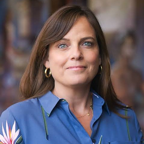 Headshot of a woman with shoulder-length brown hair, blue eyes, gold hoop earrings, and a blue button-down shirt.