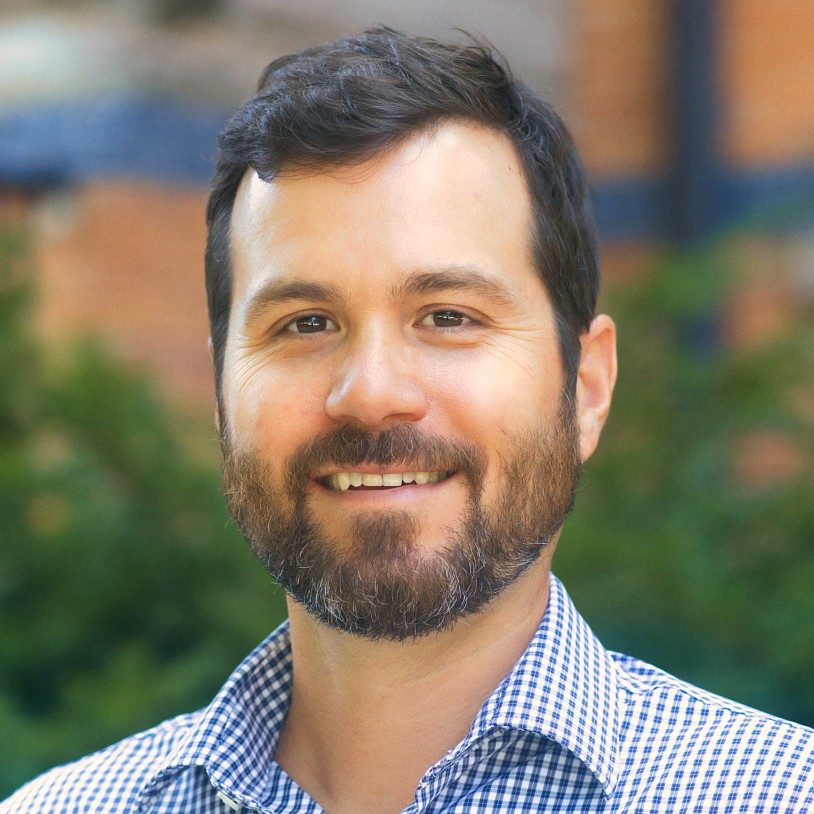 Headshot of a smiling man with dark hair and a beard wearing a blue-and-white checkered shirt, with greenery and a building blurred in the background.