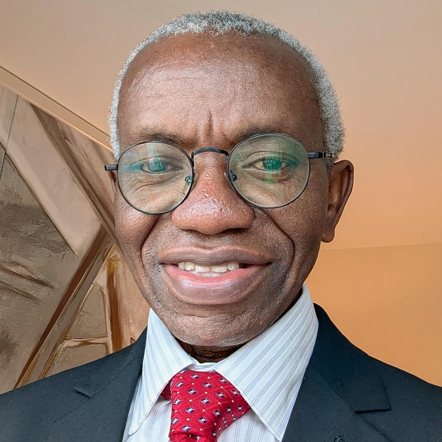 Headshot of a smiling man with short graying hair and black round-frame glasses wearing a dark suit jacket, light striped dress shirt, and a red patterned tie.