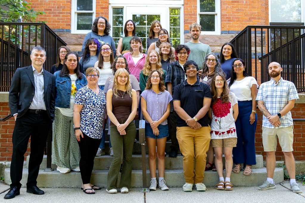 A group of more than 20 people pose together on the steps outside a brick building.