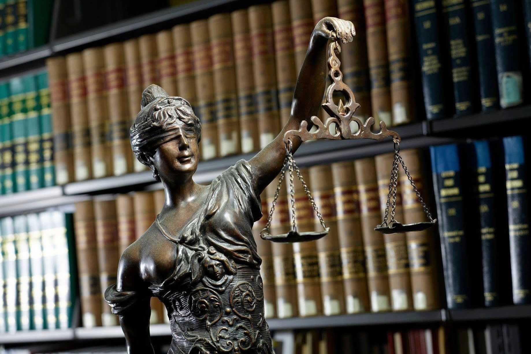 A bronze statue of Lady Justice holding scales stands in front of shelves filled with law books.