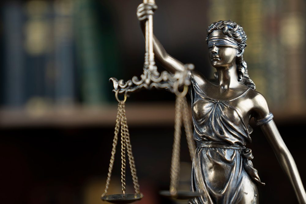 Blindfolded Lady Justice statue holding scales with blurred law books in the background.