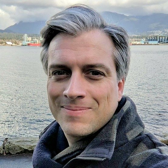 Outdoor selfie of a man with gray hair wearing a dark jacket and scarf, with mountains and an industrial waterfront in the background.