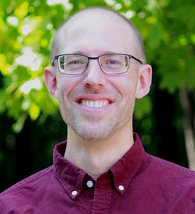 Portrait of a smiling man with glasses wearing a burgundy button-down shirt, photographed outdoors against a leafy green background.