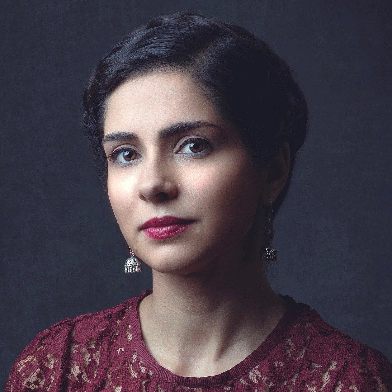 Portrait of a woman with dark hair styled back, wearing silver earrings and a burgundy lace top against a dark background.