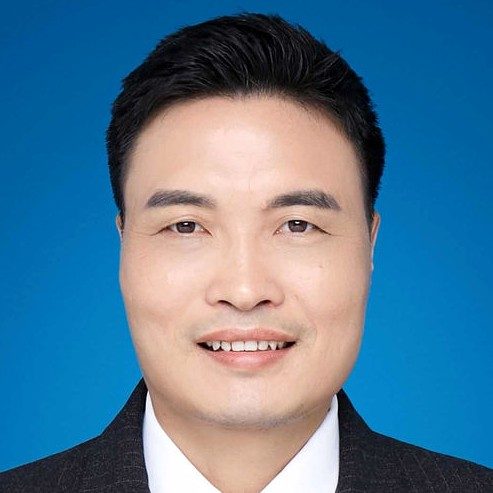 Headshot of a smiling man with short dark hair wearing a dark suit and white shirt against a blue background.