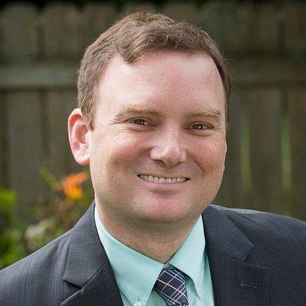 Portrait of a smiling man with short brown hair wearing a dark suit jacket, light green shirt, and patterned tie, photographed outdoors.