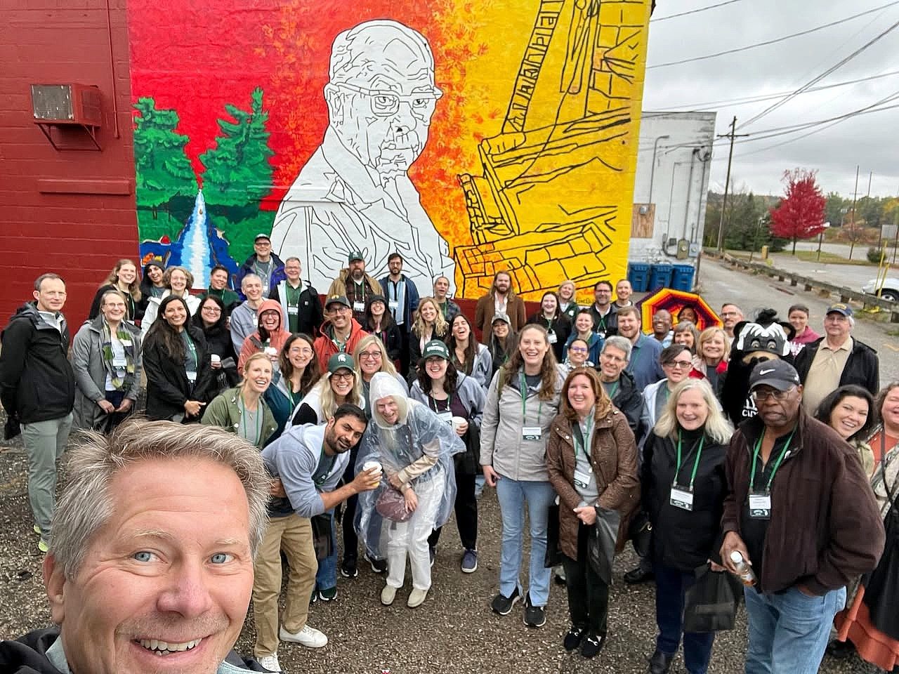 A large group of people pose for a photo in front of the completed mural featuring an outlined portrait of an older man with trees and a river on one side. The group smiles and waves, gathered outdoors on a cloudy day.