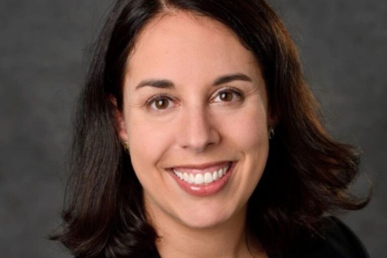 A woman with shoulder length black hair, brown eyes, and light skin smiling and wearing a black jacket over a mosaic print purple and white top.