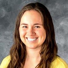 Headshot of a smiling woman with long brown hair wearing a yellow top against a gray background.