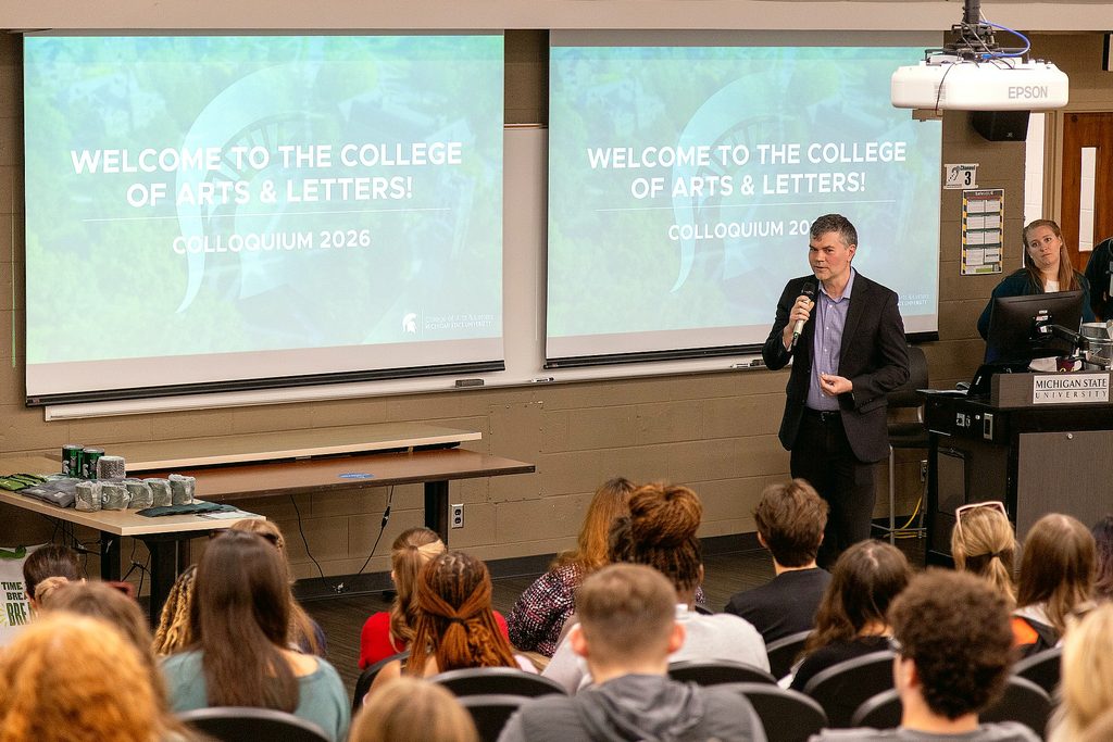 A man speaks into a microphone before a seated audience during the College of Arts & Letters Colloquium 2026, with welcome slides projected behind him.