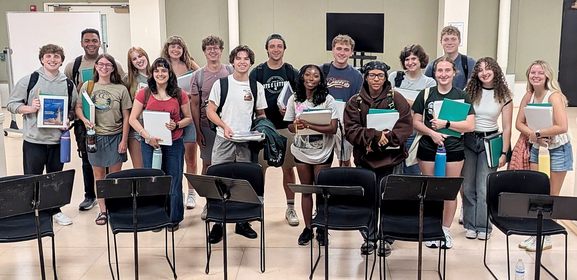 Cast members of “Discovery: A New American Musical” pose together in a rehearsal room, holding scripts and binders behind a row of chairs and music stands.