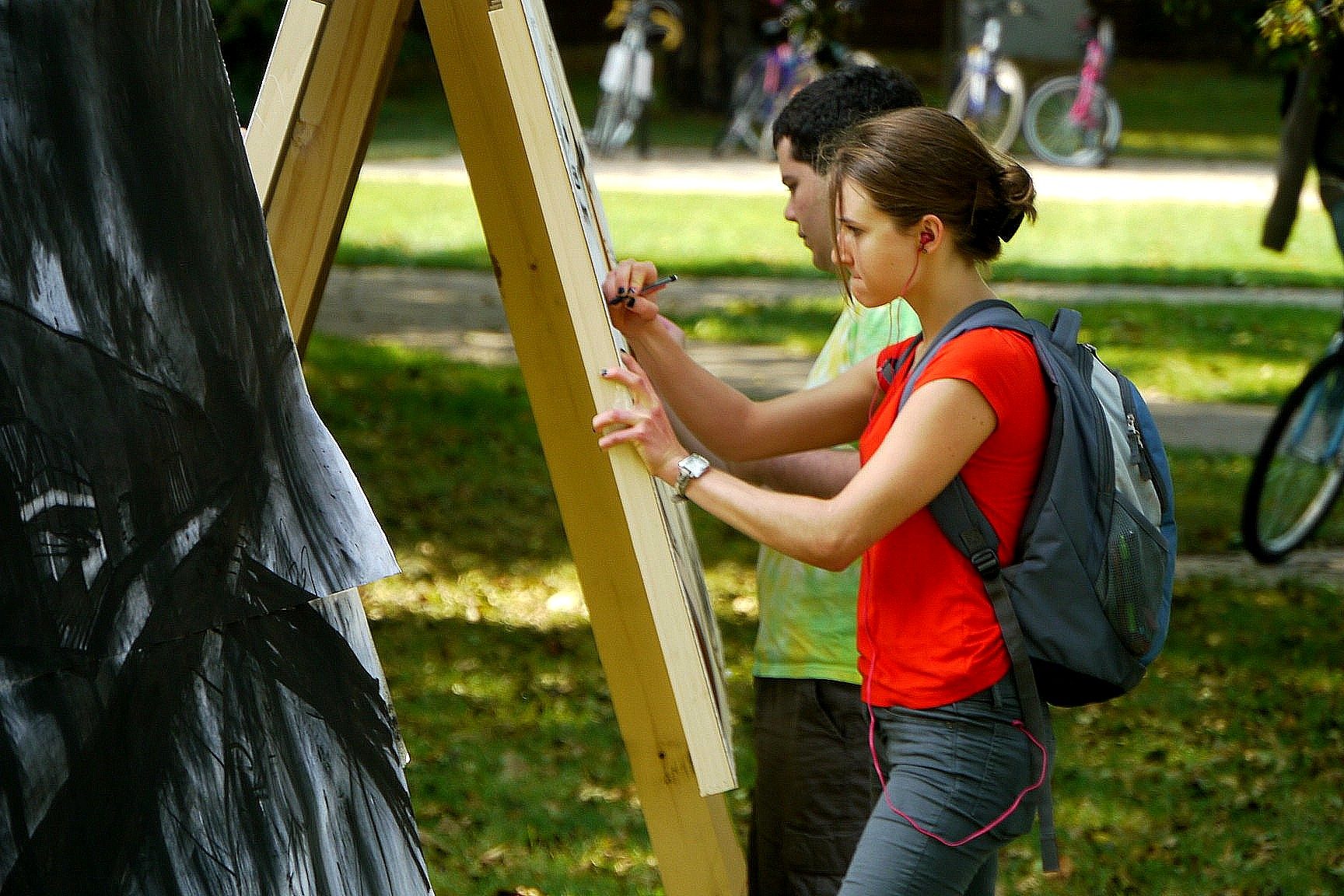 A student wearing a red shirt and backpack works on a large black-and-white drawing mounted on an outdoor easel during the MSU Drawing Marathon.