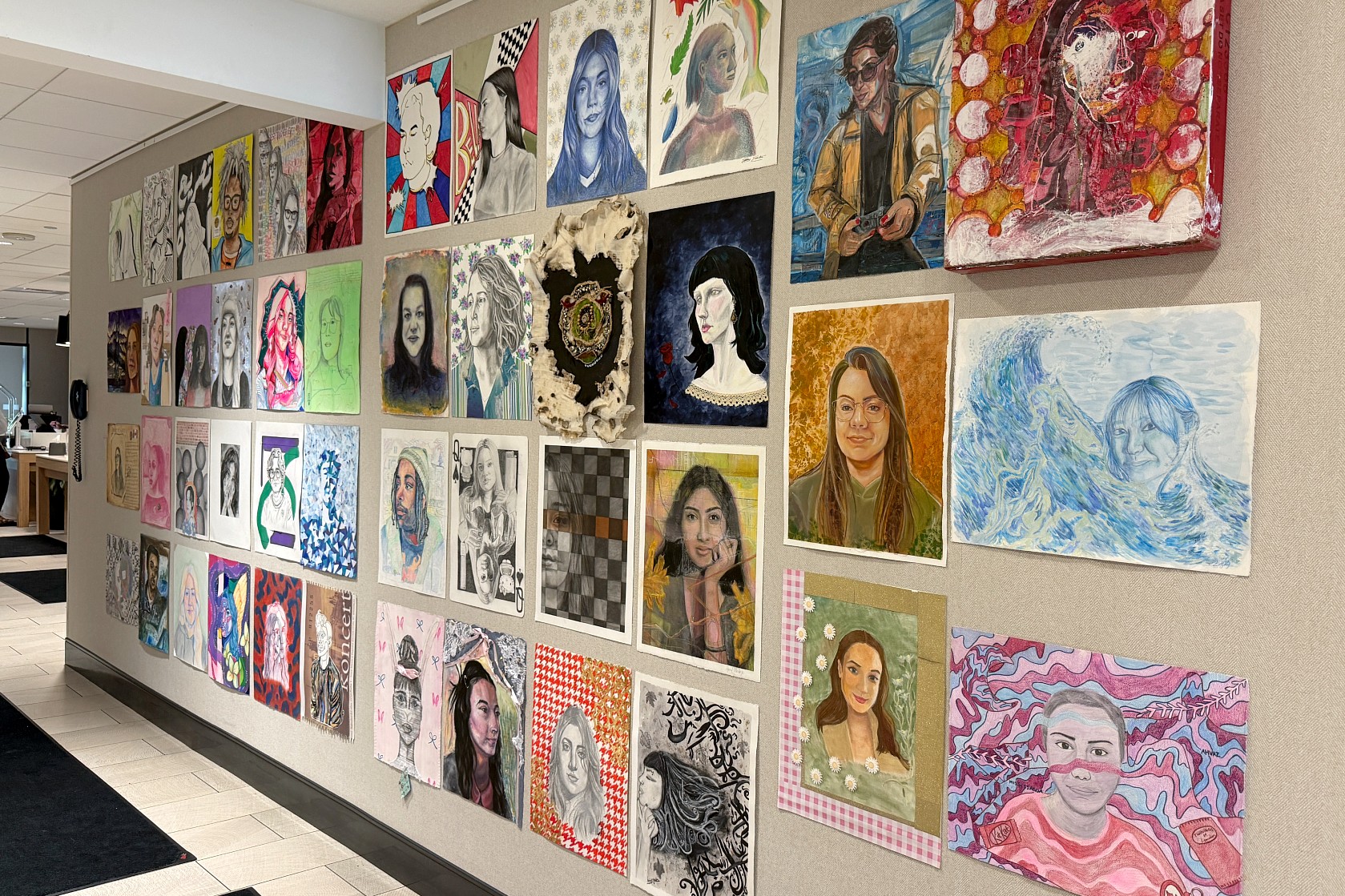 An angled view of the “Faces and Traces: A Cross-Border Portrait Project” exhibition shows dozens of student-created portraits in a range of artistic styles and media displayed together on a wall.