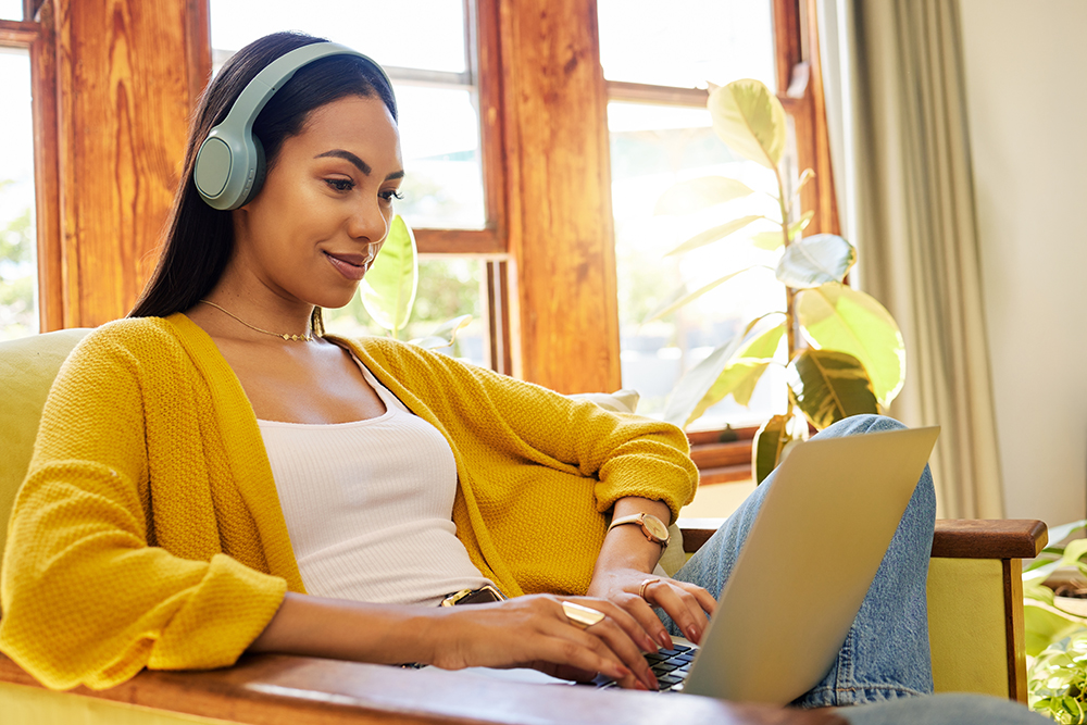 A woman wearing headphones and a yellow sweater sits in a comfortable chair with a laptop in her lap.