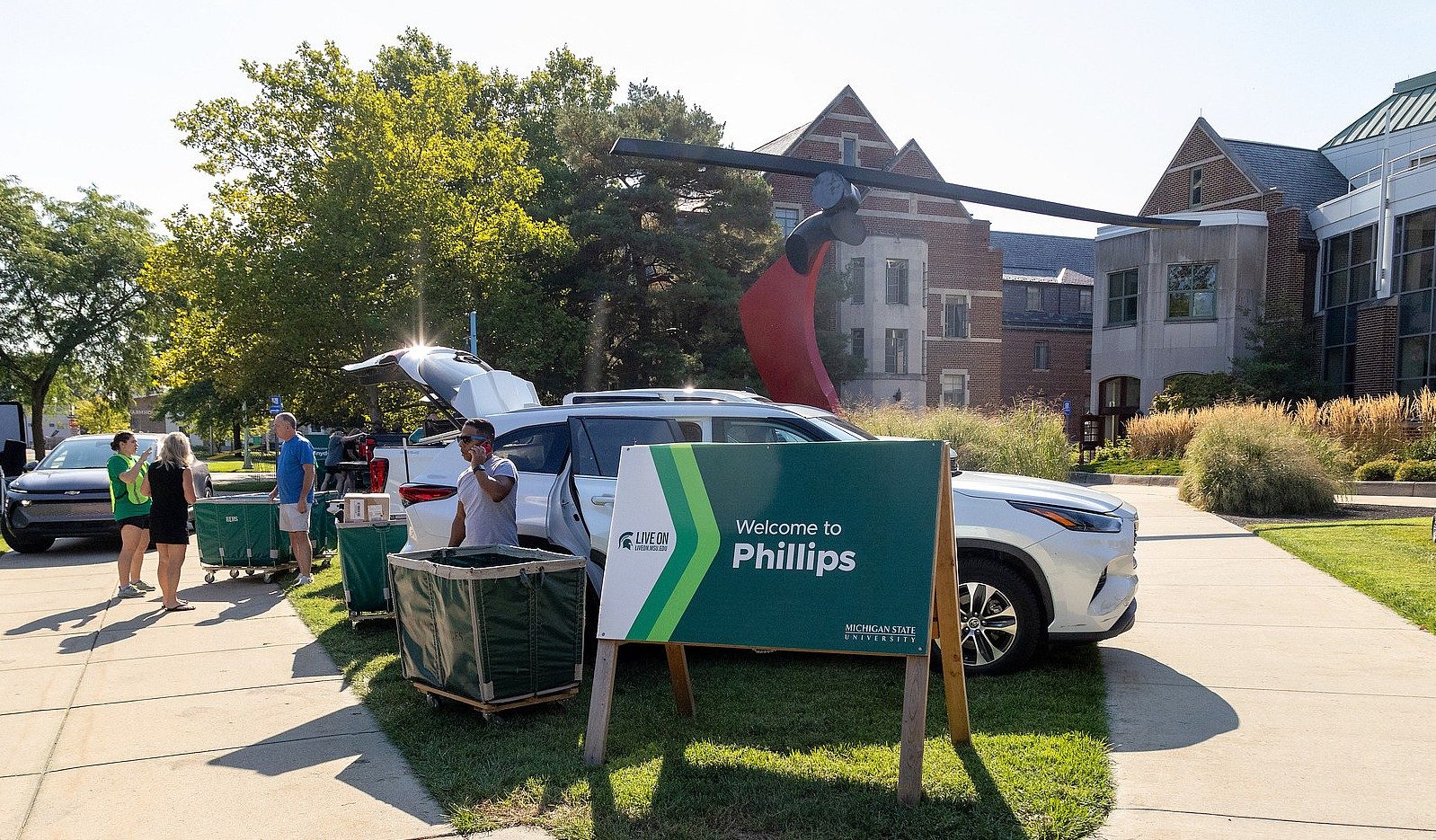 People unload vehicles and move belongings outside Phillips Hall during move-in, with a Michigan State University welcome sign in the foreground.
