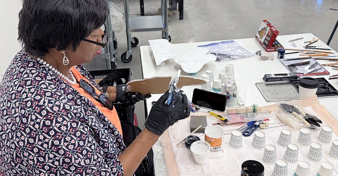 A woman wearing gloves applies paint to a piece of cardboard at a worktable covered with art supplies, brushes, tools, and paper cups.