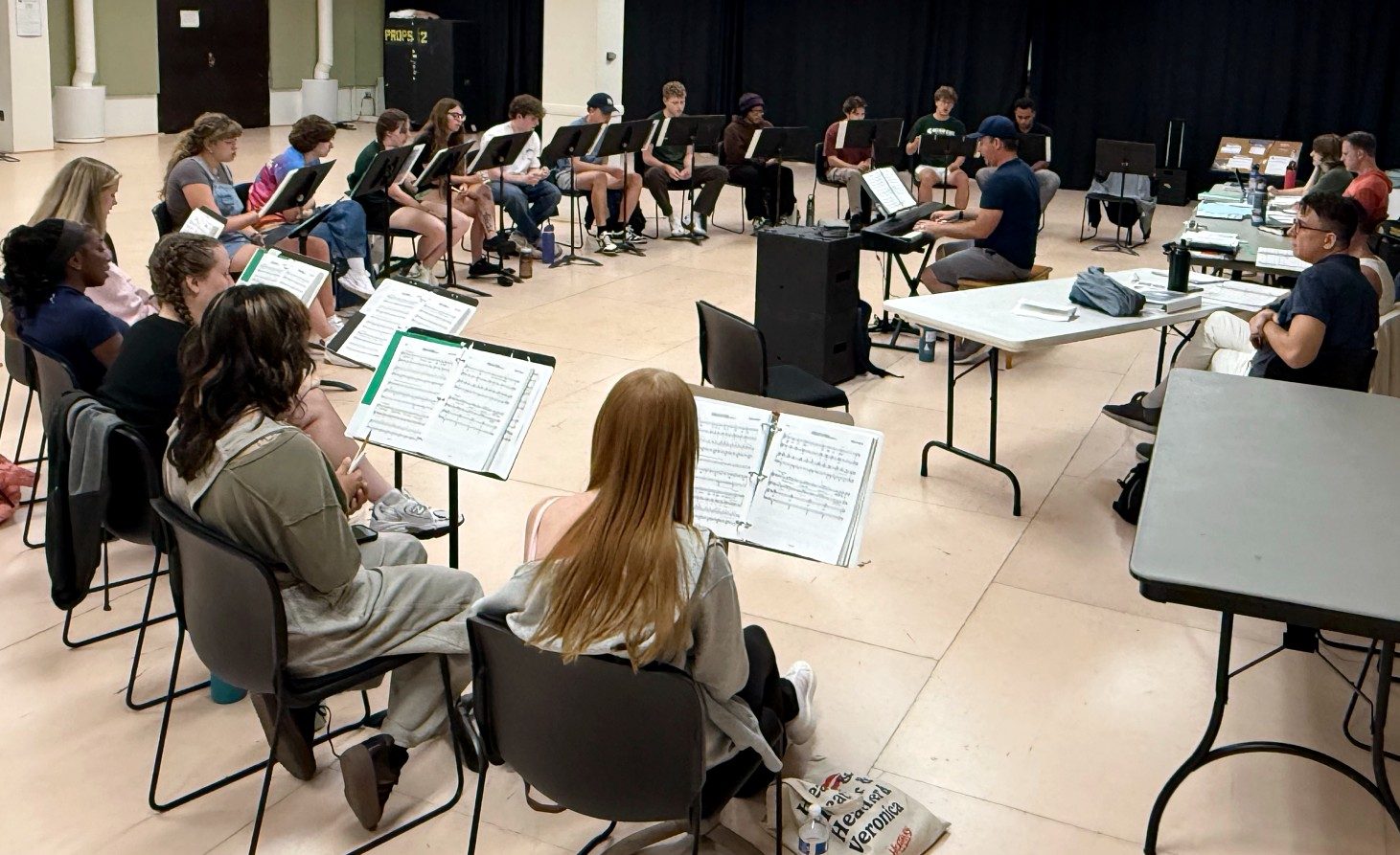 Cast members of “Discovery: A New American Musical” sit in a circle with scripts and sheet music during a rehearsal as a pianist plays at the center of the room.