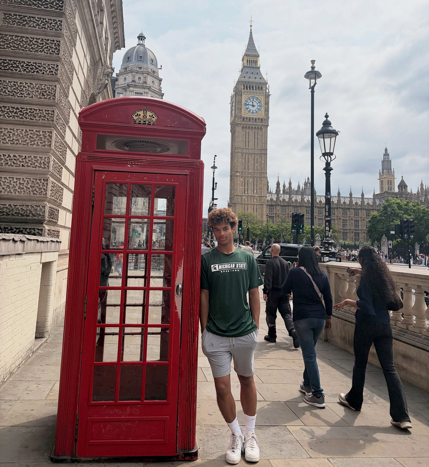 Michigan State University student Anthony Wilson stands beside a traditional red British telephone booth, wearing a green MSU Spartans shirt, with Big Ben and the Palace of Westminster in the background in London, England.