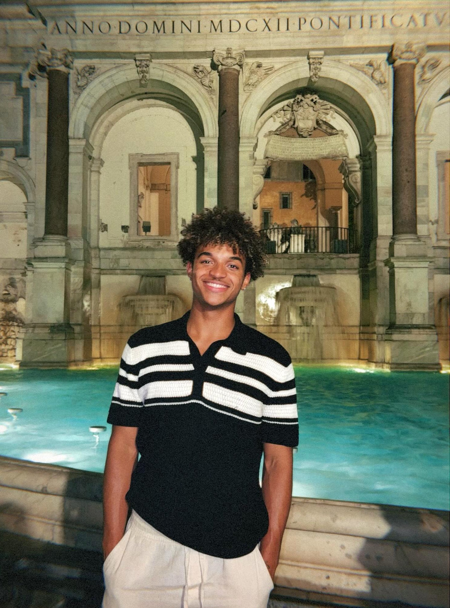 Anthony Wilson smiles in front of an illuminated fountain with turquoise water, ornate stone arches, and columns in the background in Rome, Italy.