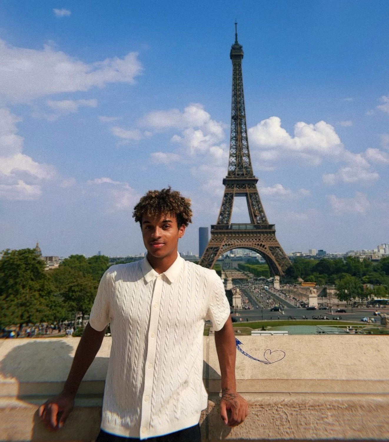 Anthony Wilson poses in a white short-sleeved shirt with the Eiffel Tower rising behind him against a partly cloudy sky in Paris, France.