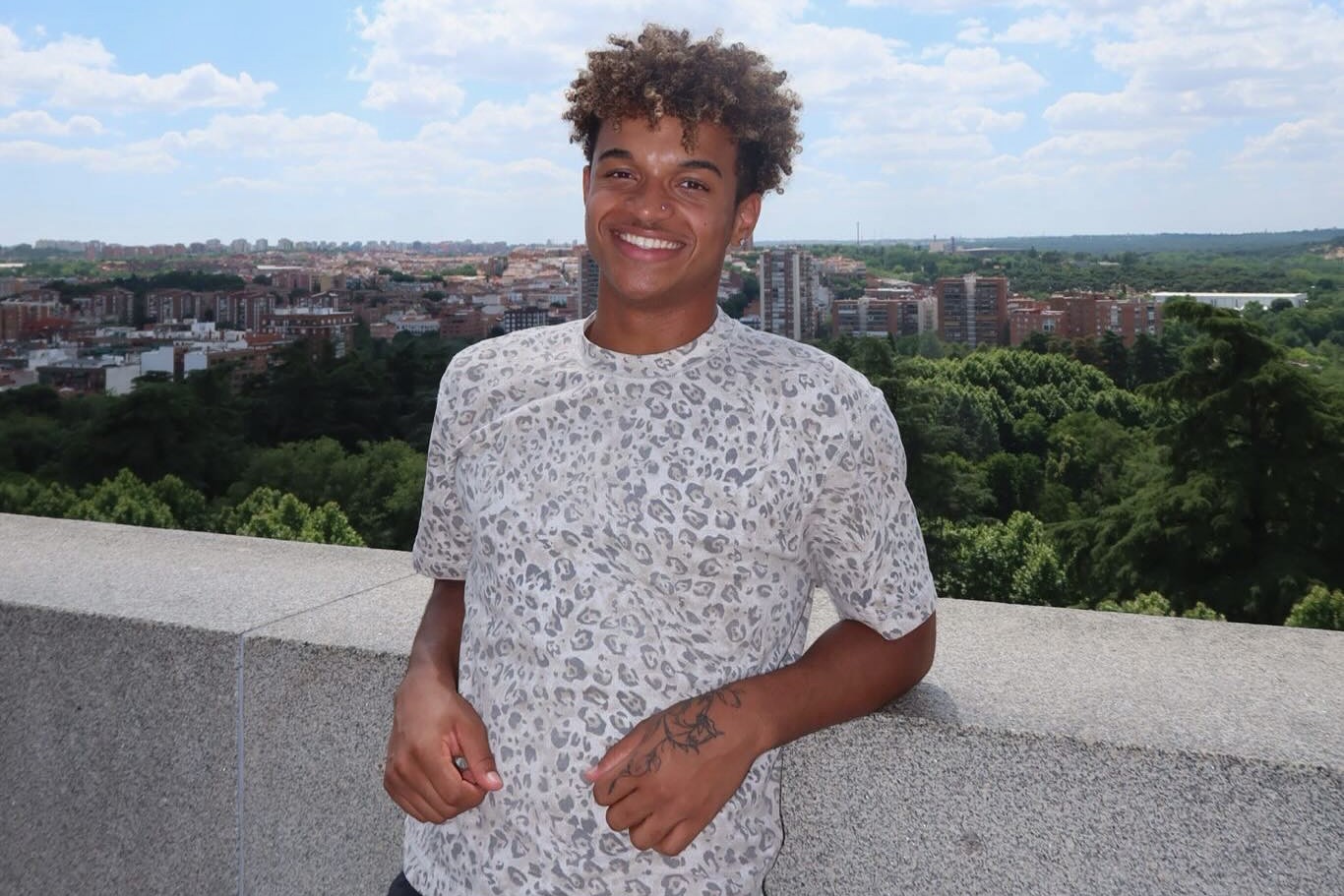Anthony Wilson smiles while leaning against a stone railing overlooking a tree-filled landscape and city buildings in Spain, wearing a white patterned shirt.
