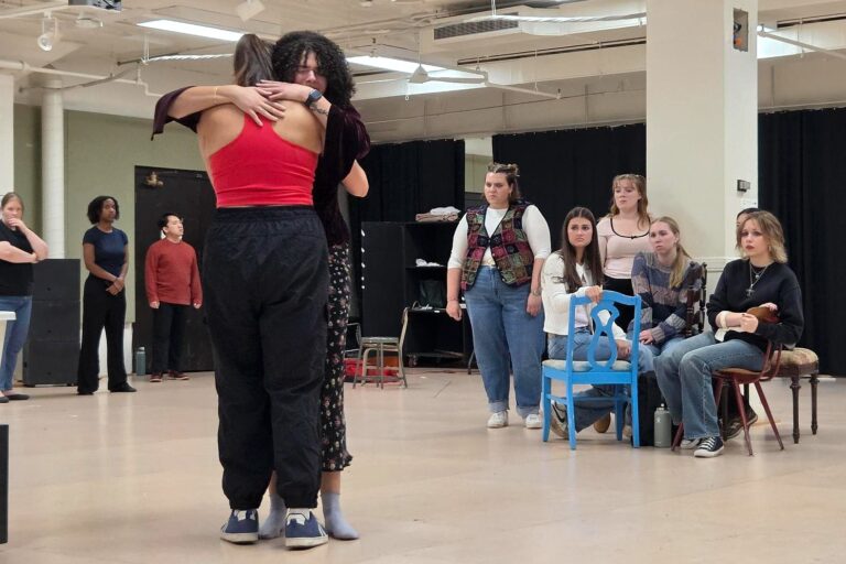 Two performers embrace during a rehearsal of MSU Theatre’s “Antigone” as fellow cast members watch from around the rehearsal space, some seated in chairs.