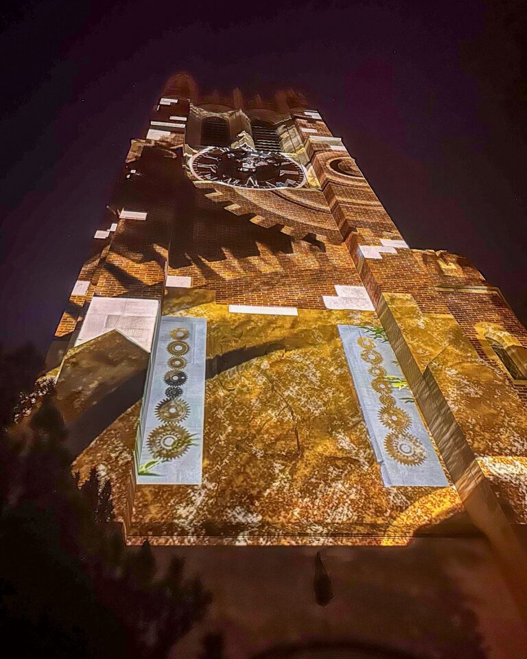 Golden and brown projections cover Beaumont Tower, featuring intricate gear designs, textured patterns, and large mechanical shapes illuminated against the dark sky.