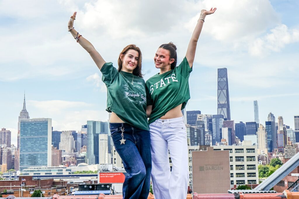 Michigan State University students Nadia Bednar and Casey Klau pose with their arms raised in front of the New York City skyline, wearing green MSU shirts.