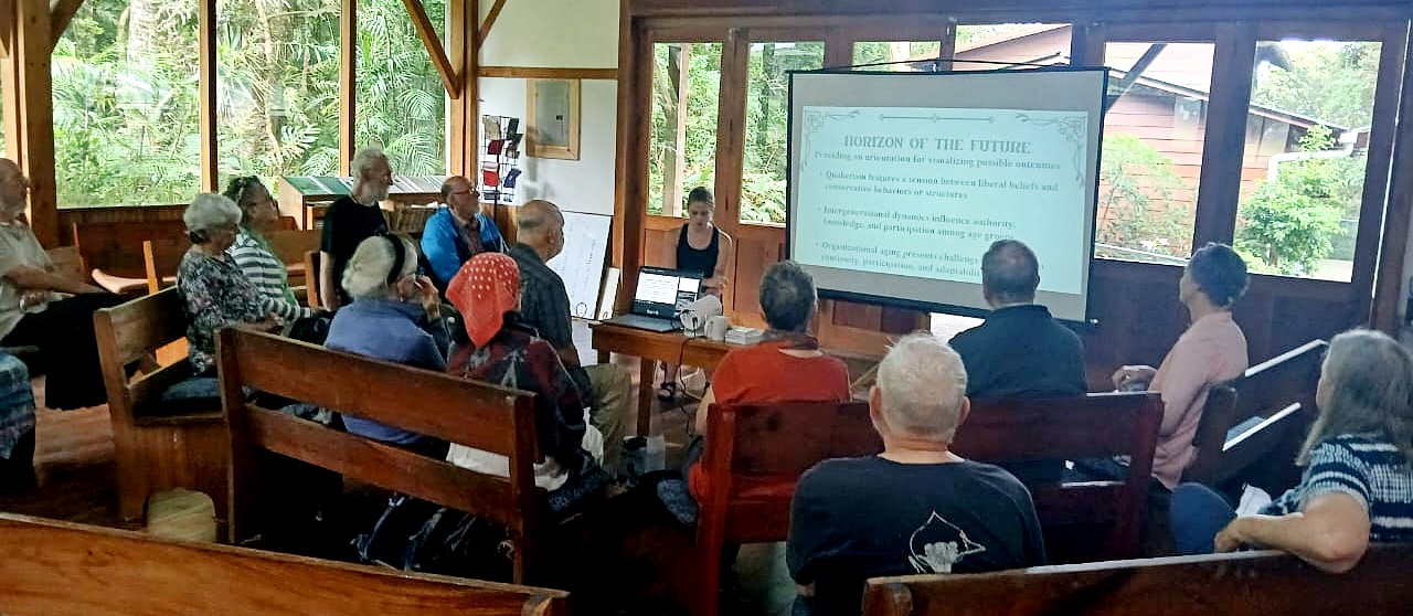 A wider view of a meeting house with large windows overlooking lush vegetation shows a group of attendees gathered on wooden benches for a presentation titled “Horizon of the Future.” A woman sits at the front beside a projected slide discussing organizational challenges and future possibilities.
