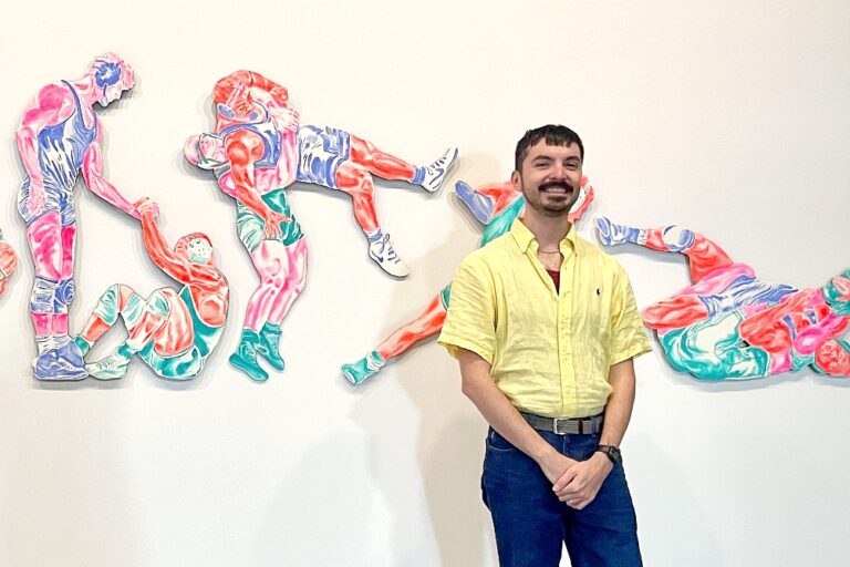 A smiling man in a yellow shirt stands beside a vibrant gallery wall installation of colorful abstract figures in motion.