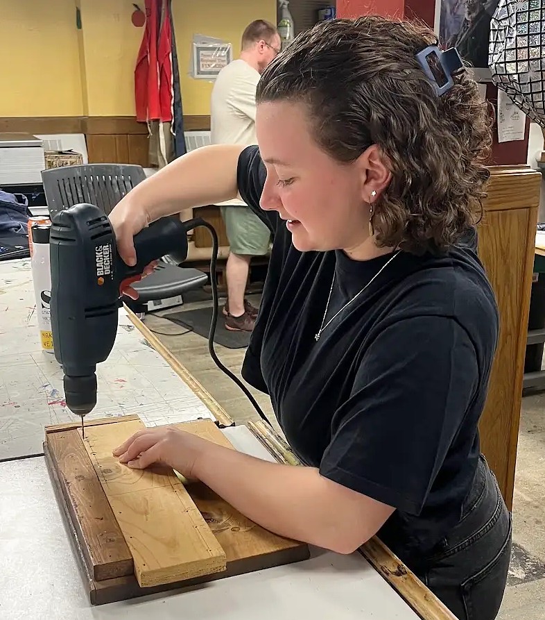 A student uses a power drill to make holes in a wooden board while working on a project in a studio.