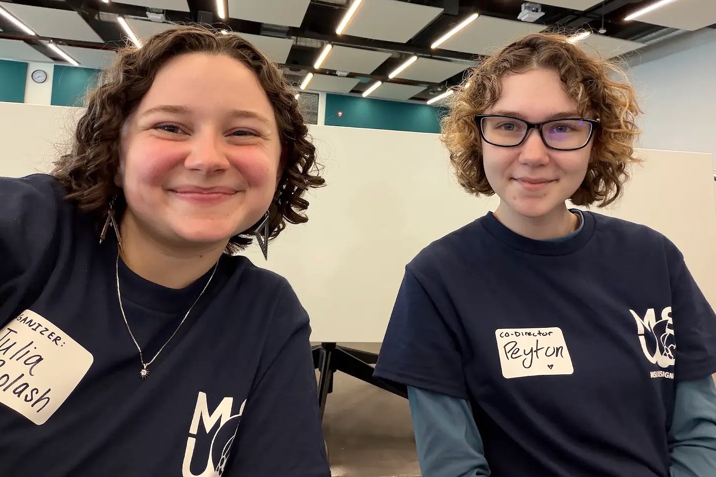 Two young adults wearing T-shirts sit side by side and smile at the camera. Both wear name tags identifying them as event organizers. They are pictured in a bright indoor workspace.