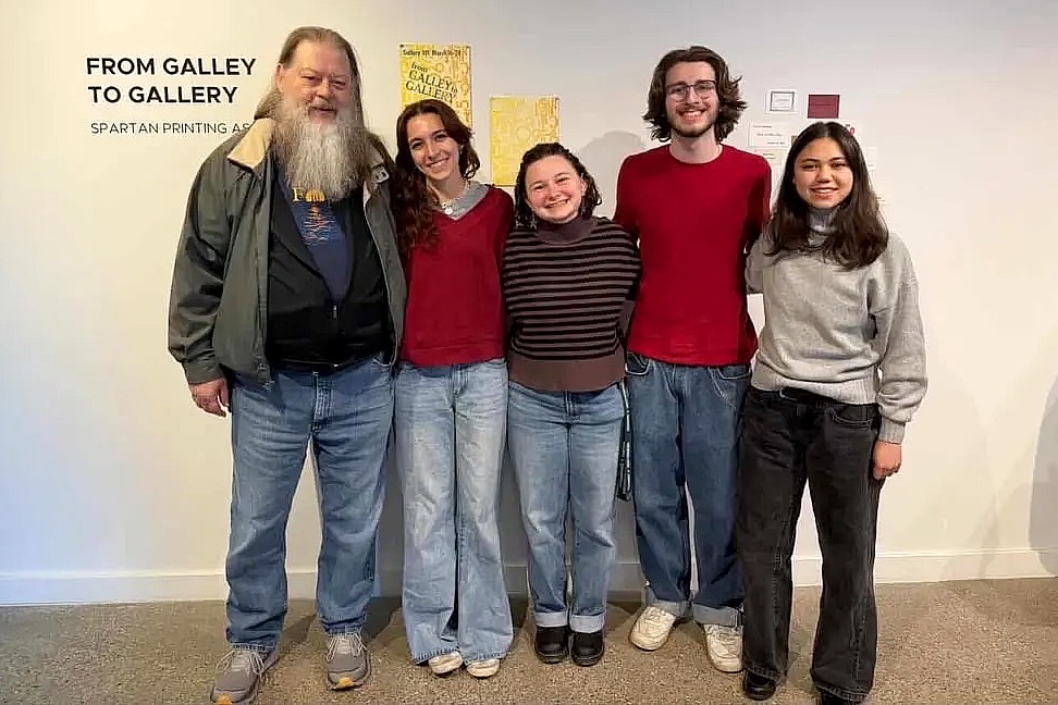 A group of five people, including an older man with a long white beard and four young adults, stand together smiling in front of an exhibition wall titled “From Galley to Gallery: Spartan Printing.” They pose for a group photo at a printing-related exhibit.