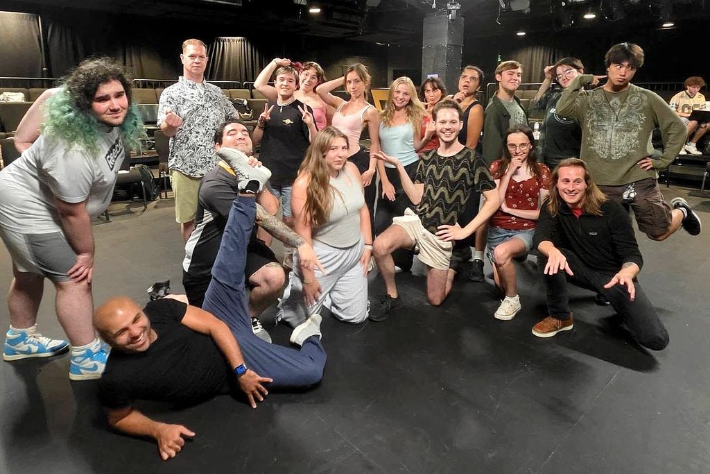 A group of people pose together indoors, many with expressive or playful gestures, while one person lies stretched out at the front. Theater seats are visible in the background.