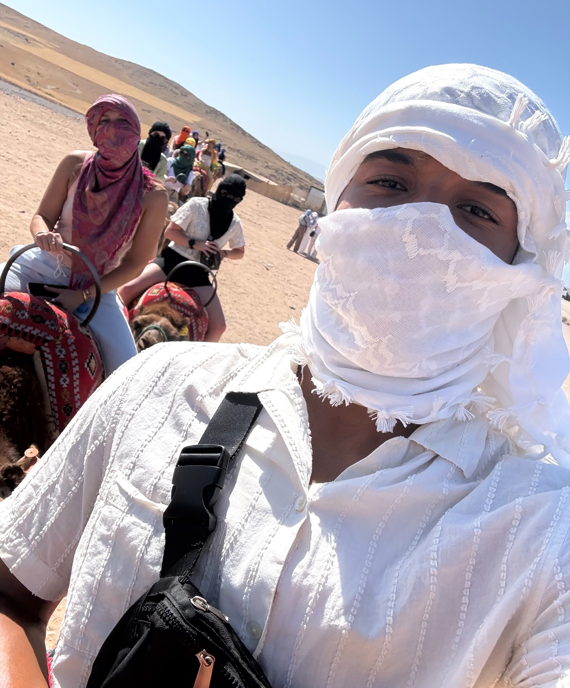 Anthony Wilson wearing a white headscarf and face covering takes a selfie while riding a camel, with other riders following behind across a sunlit, rocky desert landscape in Morocco.