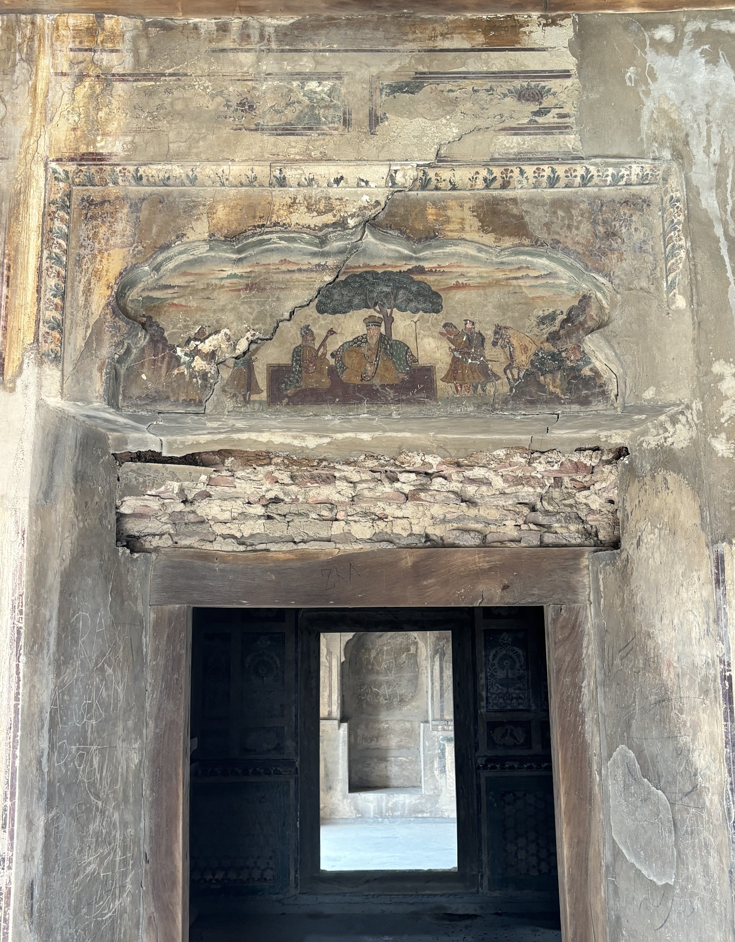 A deteriorating wall painting depicting figures gathered beneath a tree above a doorway in a historic building. Cracked plaster and exposed brick reveal the structure’s age.